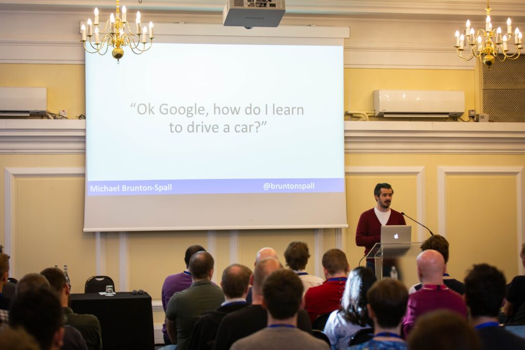 Michael Brunton-Spall talking to the audience in front of a screen about voice assistance which says: "Ok Google, how do I learn to drive a car?"