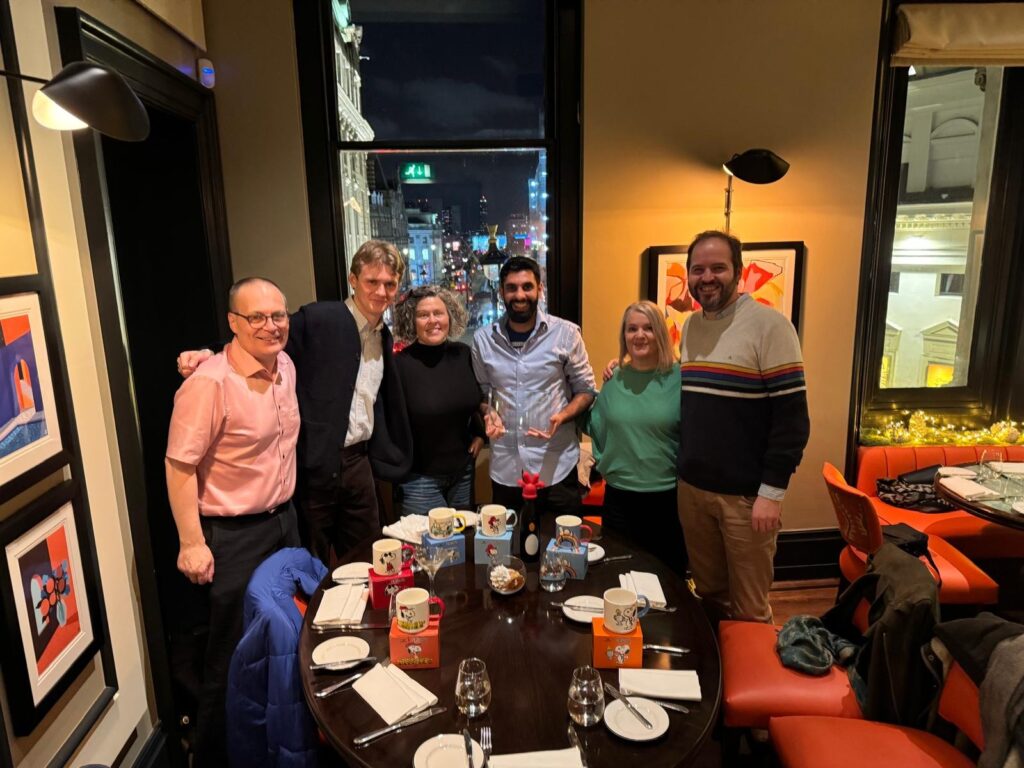 Six people stand close together and smile for a group photo around a set dining table in a restaurant with large windows overlooking a city at night.
