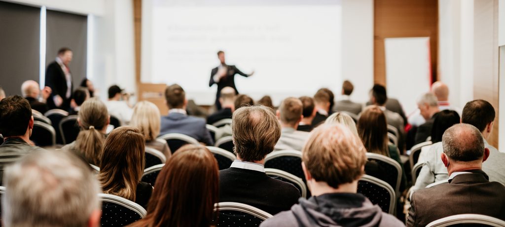 A large audience sits in a conference hall, facing a large screen displaying a presentation. The room is filled with people attending a business event or seminar.